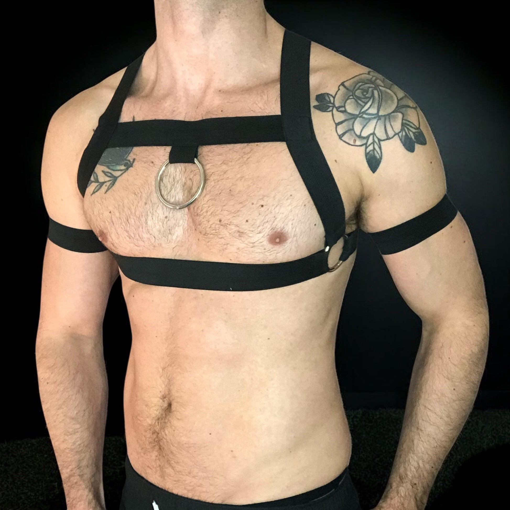 Man wearing a custom-fit black body harness with metal ring and matching bicep bands by CheekyBoyHarness. Festival and clubwear style, handmade to measure