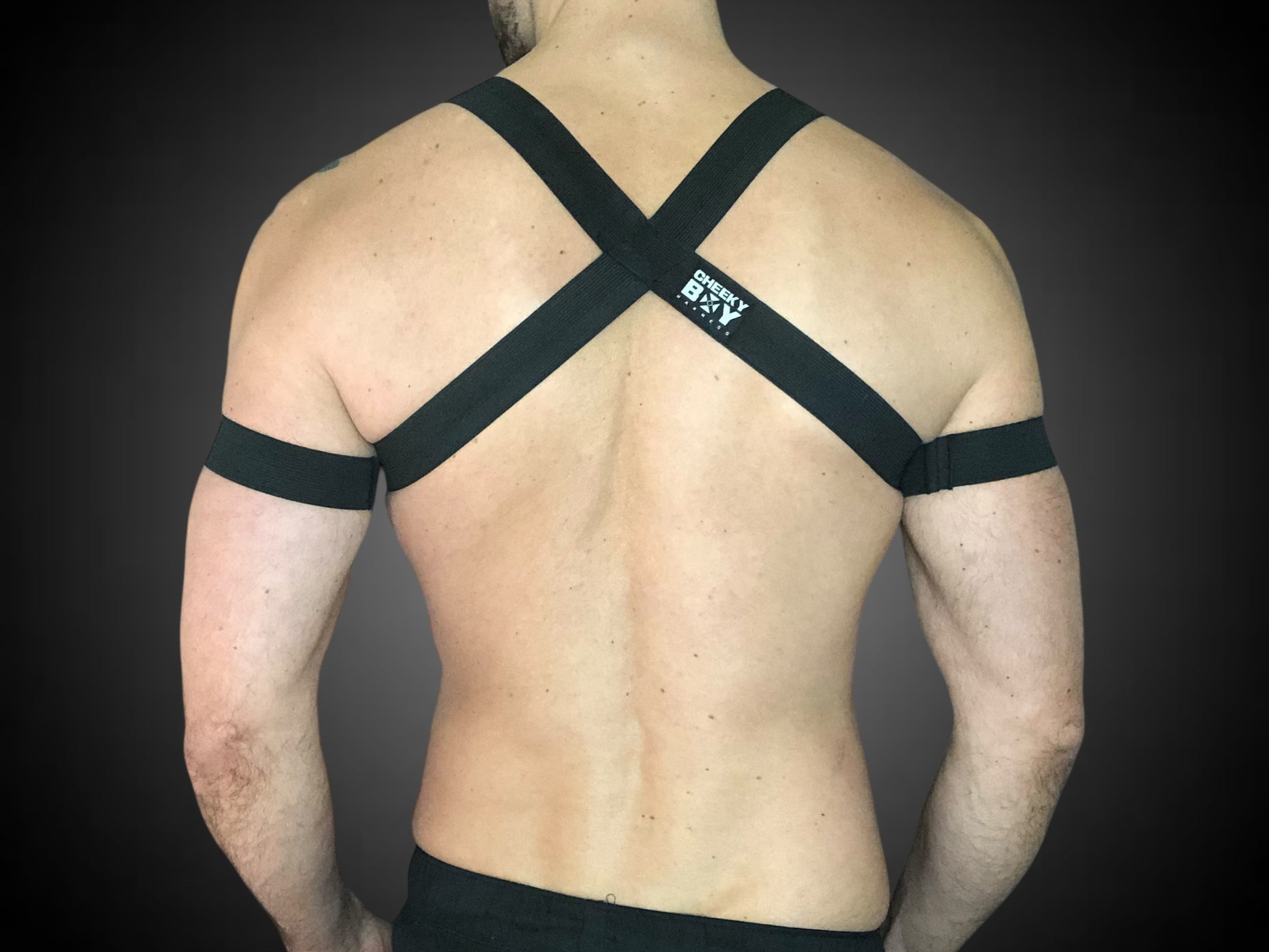 Man wearing a custom-fit black body harness with metal ring and matching bicep bands by CheekyBoyHarness. Festival and clubwear style, handmade to measure