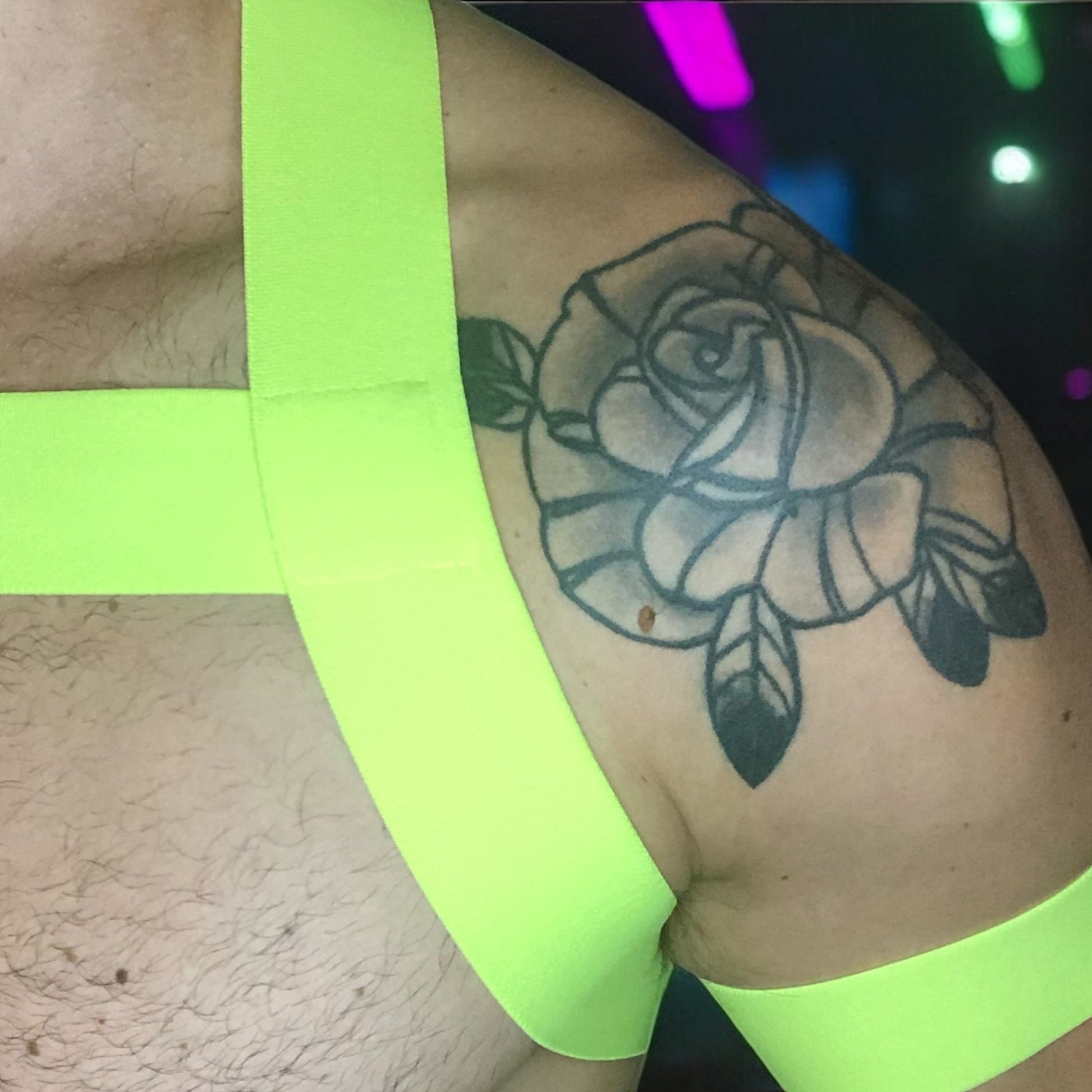 Model wearing a custom-made neon yellow and pink elastic chest harness with matching jockstrap and bicep bands by Cheeky Boy Harness, photographed in a glowing club setting. Made-to-measure for a perfect fit, ideal for festivals, clubwear, and pride events