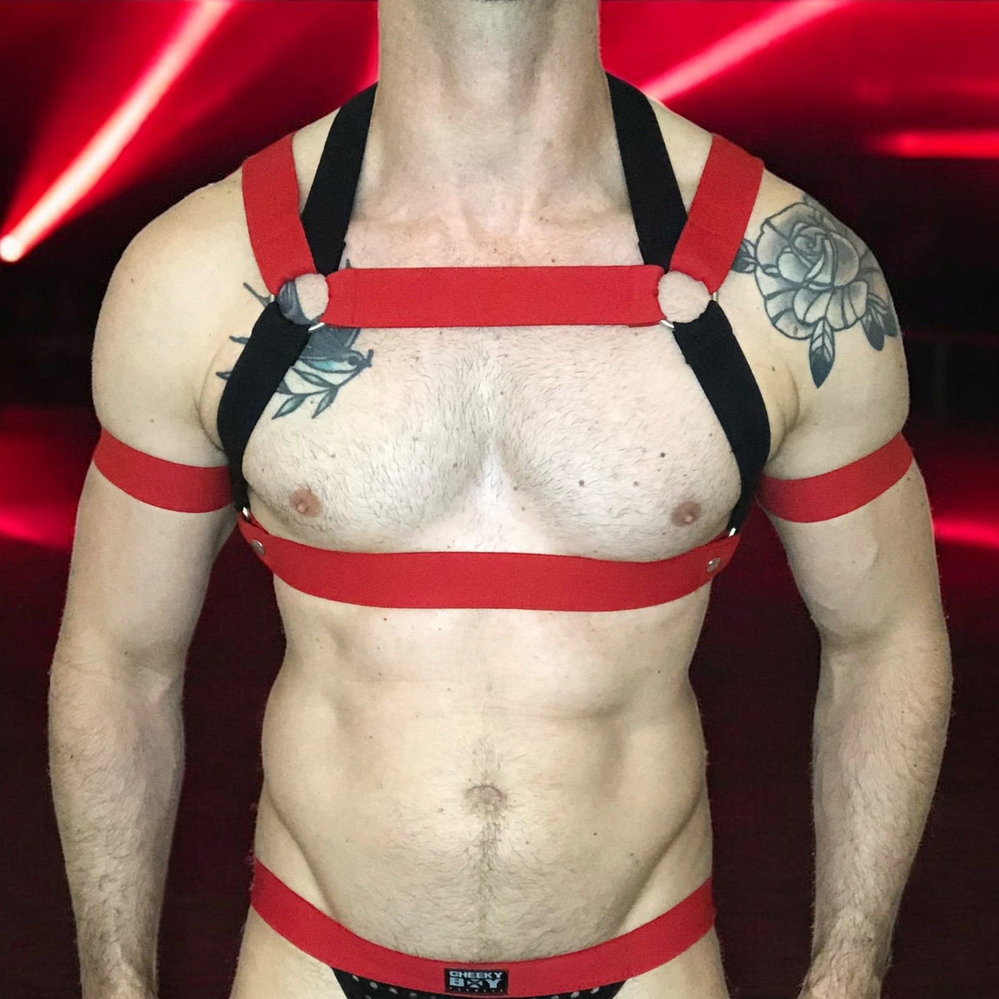 Model wearing a custom-made red and black elastic chest harness with metal rings and matching jockstrap by Cheeky Boy Harness. Includes red bicep bands. Designed for clubwear, raves, fetish events, and festivals