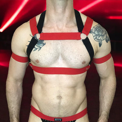 Model wearing a custom-made red and black elastic chest harness with metal rings and matching jockstrap by Cheeky Boy Harness. Includes red bicep bands. Designed for clubwear, raves, fetish events, and festivals