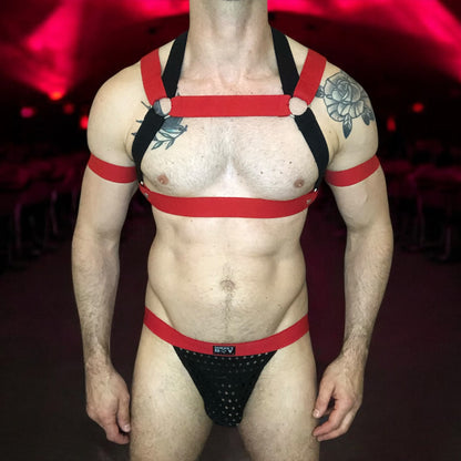 Model wearing a custom-made red and black elastic chest harness with metal rings and matching jockstrap by Cheeky Boy Harness. Includes red bicep bands. Designed for clubwear, raves, fetish events, and festivals