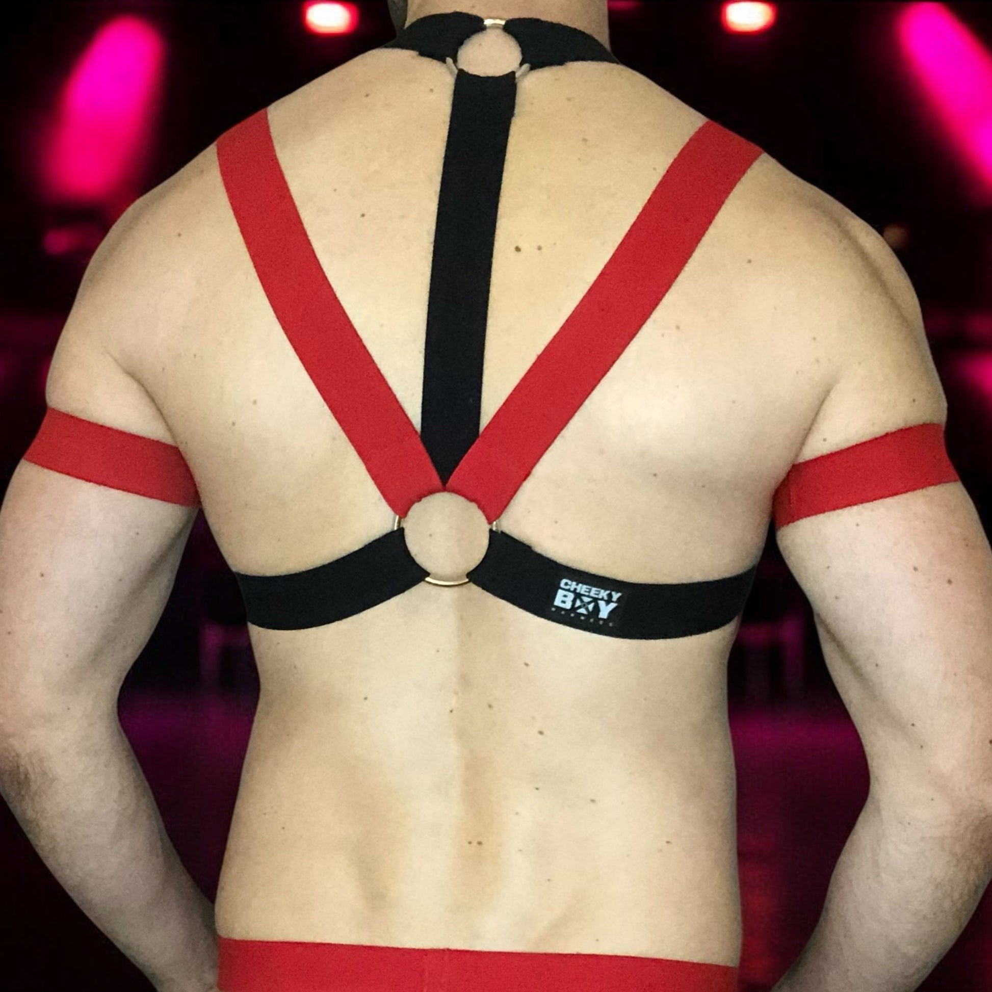 Model wearing a custom-made red and black elastic chest harness with metal rings and matching jockstrap by Cheeky Boy Harness. Includes red bicep bands. Designed for clubwear, raves, fetish events, and festivals