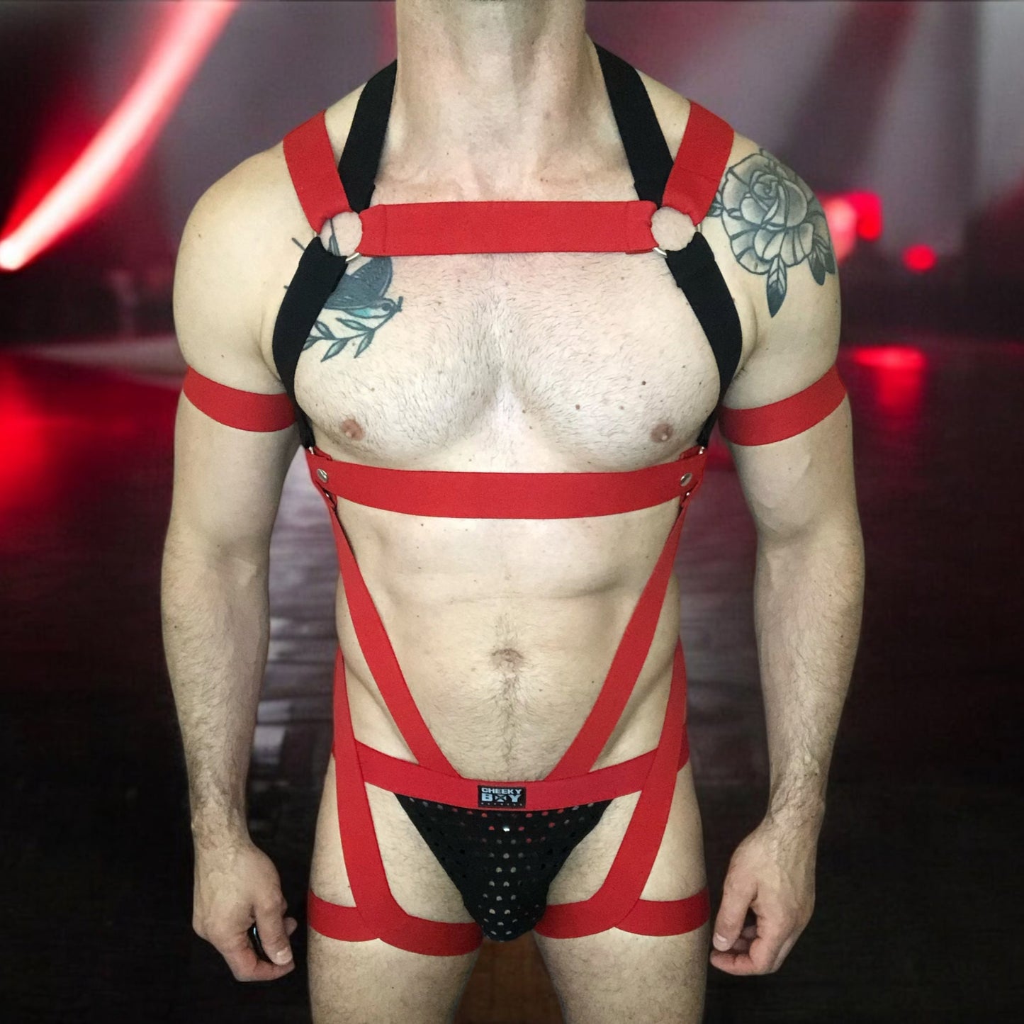 Model wearing a custom-made red and black elastic chest harness with metal rings and matching jockstrap by Cheeky Boy Harness. Includes red bicep bands. Designed for clubwear, raves, fetish events, and festivals
