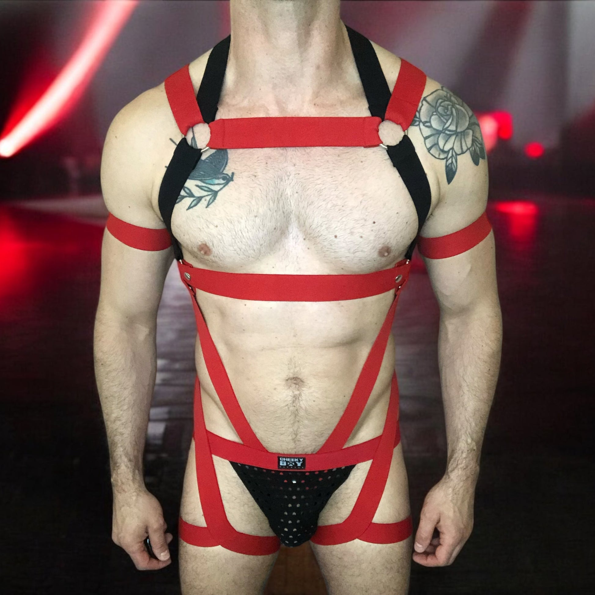 Model wearing a custom-made red and black elastic chest harness with metal rings and matching jockstrap by Cheeky Boy Harness. Includes red bicep bands. Designed for clubwear, raves, fetish events, and festivals