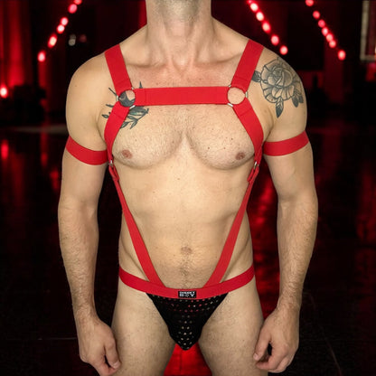 A model wearing a made-to-measure red elastic body harness by CheekyBoyHarness with silver metal rings across the chest and torso. The outfit includes matching red armbands and leg straps, styled with a black mesh jockstrap. Photographed in a red-lit club hallway, perfect for gay festival or nightlife fashion