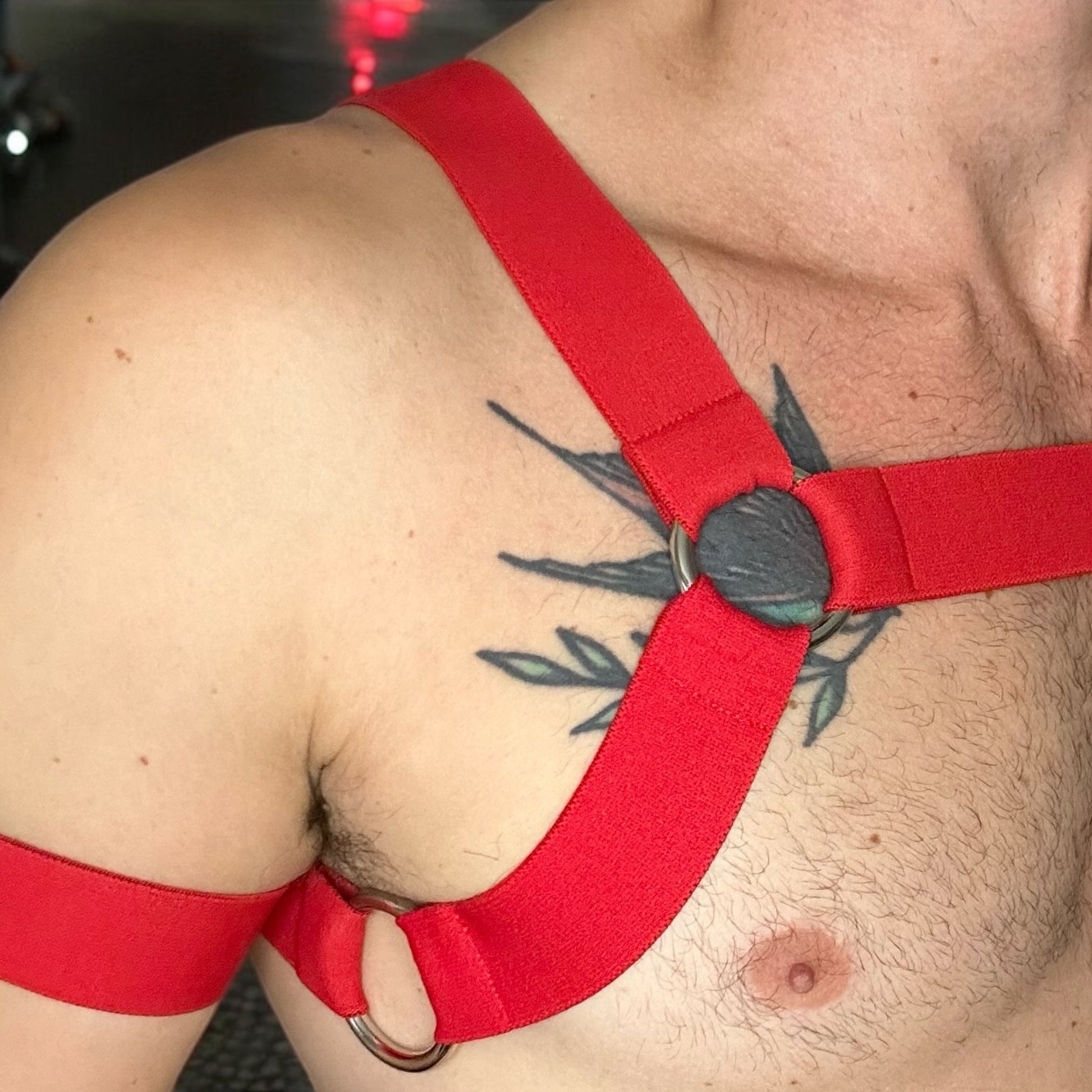 A model wearing a made-to-measure red elastic body harness by CheekyBoyHarness with silver metal rings across the chest and torso. The outfit includes matching red armbands and leg straps, styled with a black mesh jockstrap. Photographed in a red-lit club hallway, perfect for gay festival or nightlife fashion