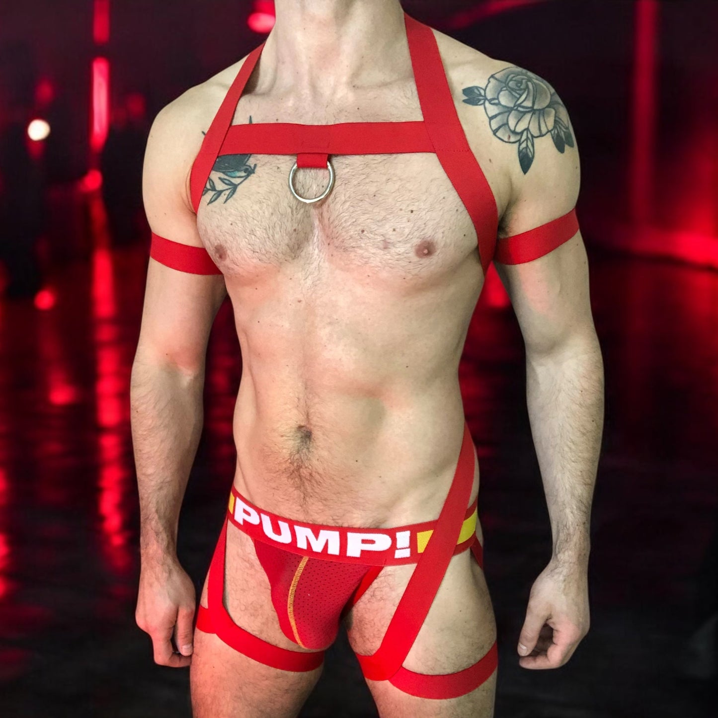 Model wearing a custom red elastic body harness by Cheeky Boy Harness, featuring a silver chest ring, matching bicep bands, and thigh straps, styled with a red mesh jockstrap.