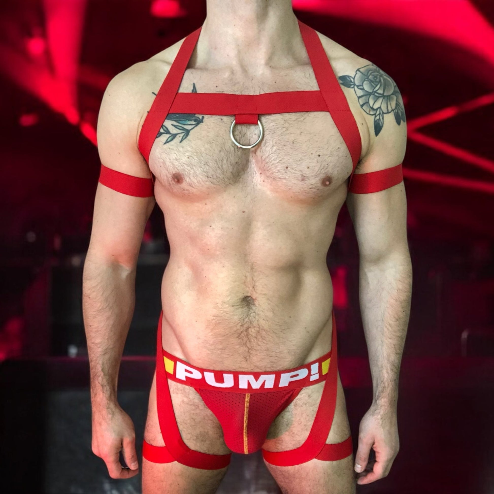 Model wearing a custom red elastic body harness by Cheeky Boy Harness, featuring a silver chest ring, matching bicep bands, and thigh straps, styled with a red mesh jockstrap.