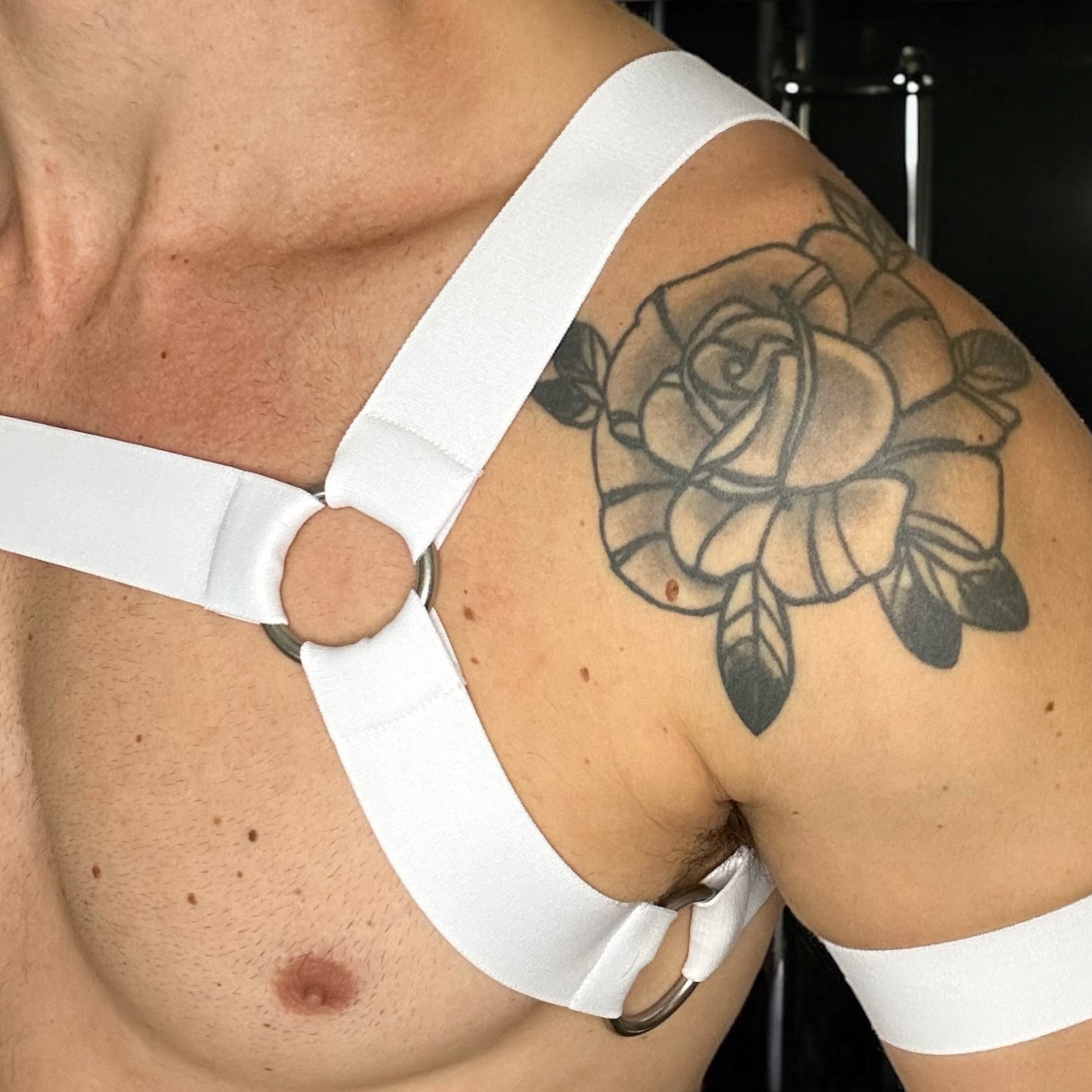 Man wearing a white elastic chest harness with 6 metal rings throughout with matching bicep bands and a white mesh jockstrap, posed in a nightclub setting, by Cheeky Boy Harness.