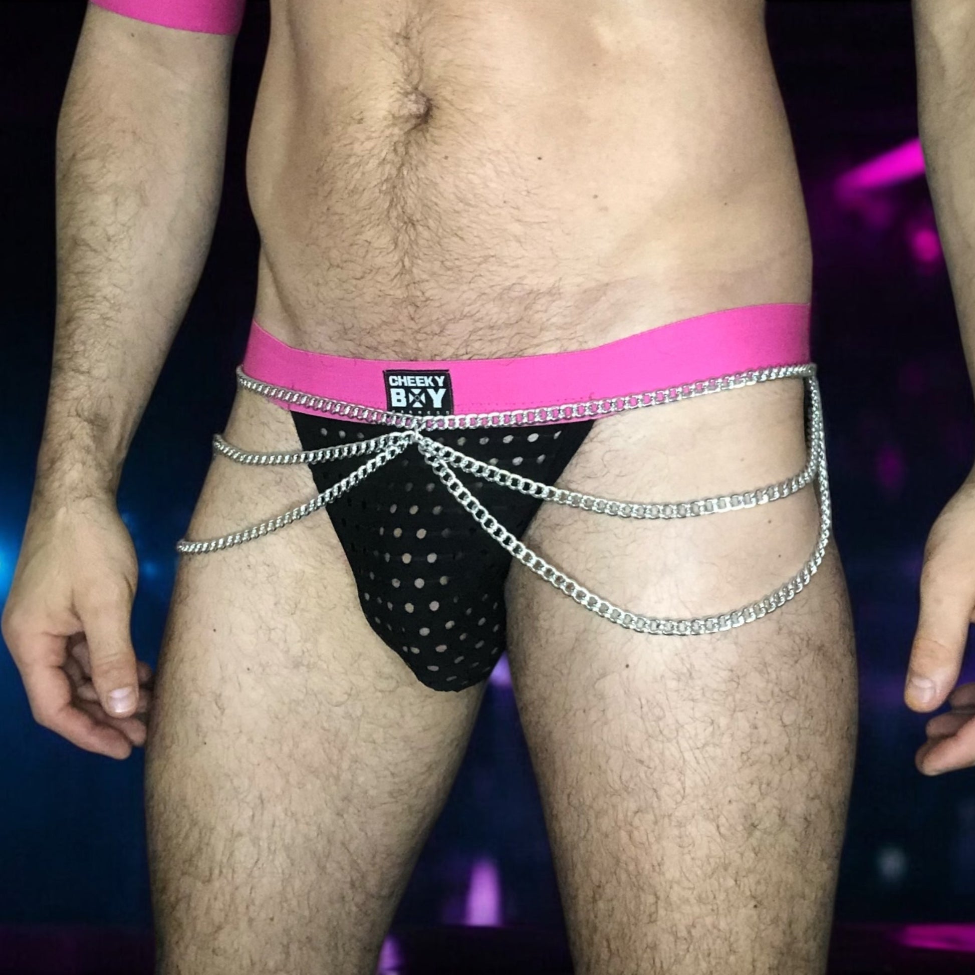 Model wearing a hot pink elastic chest harness with silver chains on chest and waistband, matching bicep bands, and a black mesh jockstrap with pink waistband by Cheeky Boy Harness.