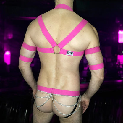 Model wearing a hot pink elastic chest harness with silver chains on chest and waistband, matching bicep bands, and a black mesh jockstrap with pink waistband by Cheeky Boy Harness.