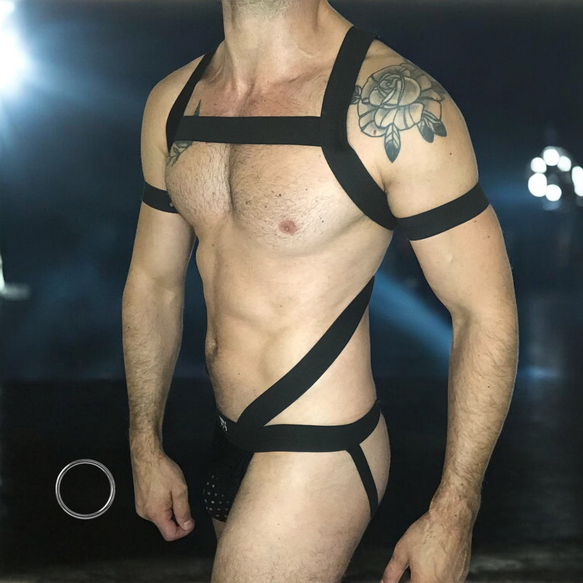 Man wearing a black elastic full body harness with metal cross over straps on back, metal chest ring, bicep bands and matching black mesh jockstrap, made by Cheeky Boy Harness