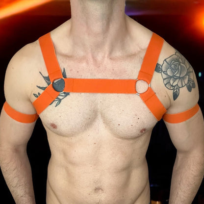 Man wearing a orange elastic chest harness with matching bicep bands and a black mesh jockstrap, posed in a nightclub setting, by Cheeky Boy Harness.