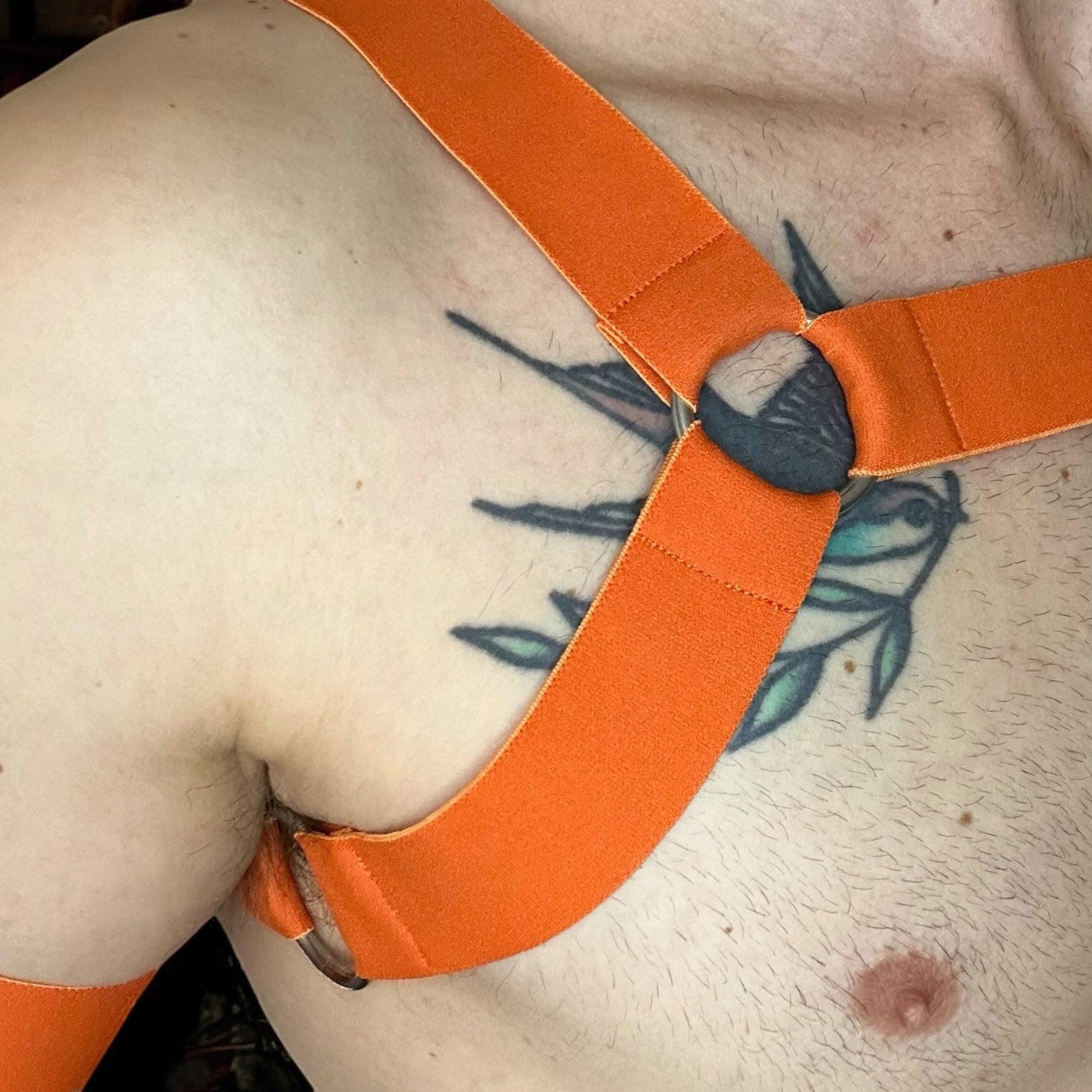 Man wearing a orange elastic chest harness with matching bicep bands and a black mesh jockstrap, posed in a nightclub setting, by Cheeky Boy Harness.