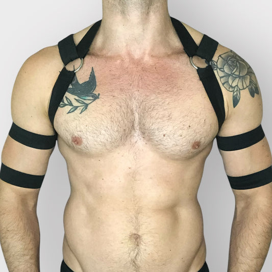 A man wearing a handmade, custom-fit black elastic full body harness with metal chain loops over chest. 4 bicep bands, and a black mesh jockstrap by Cheeky Boy Harness,