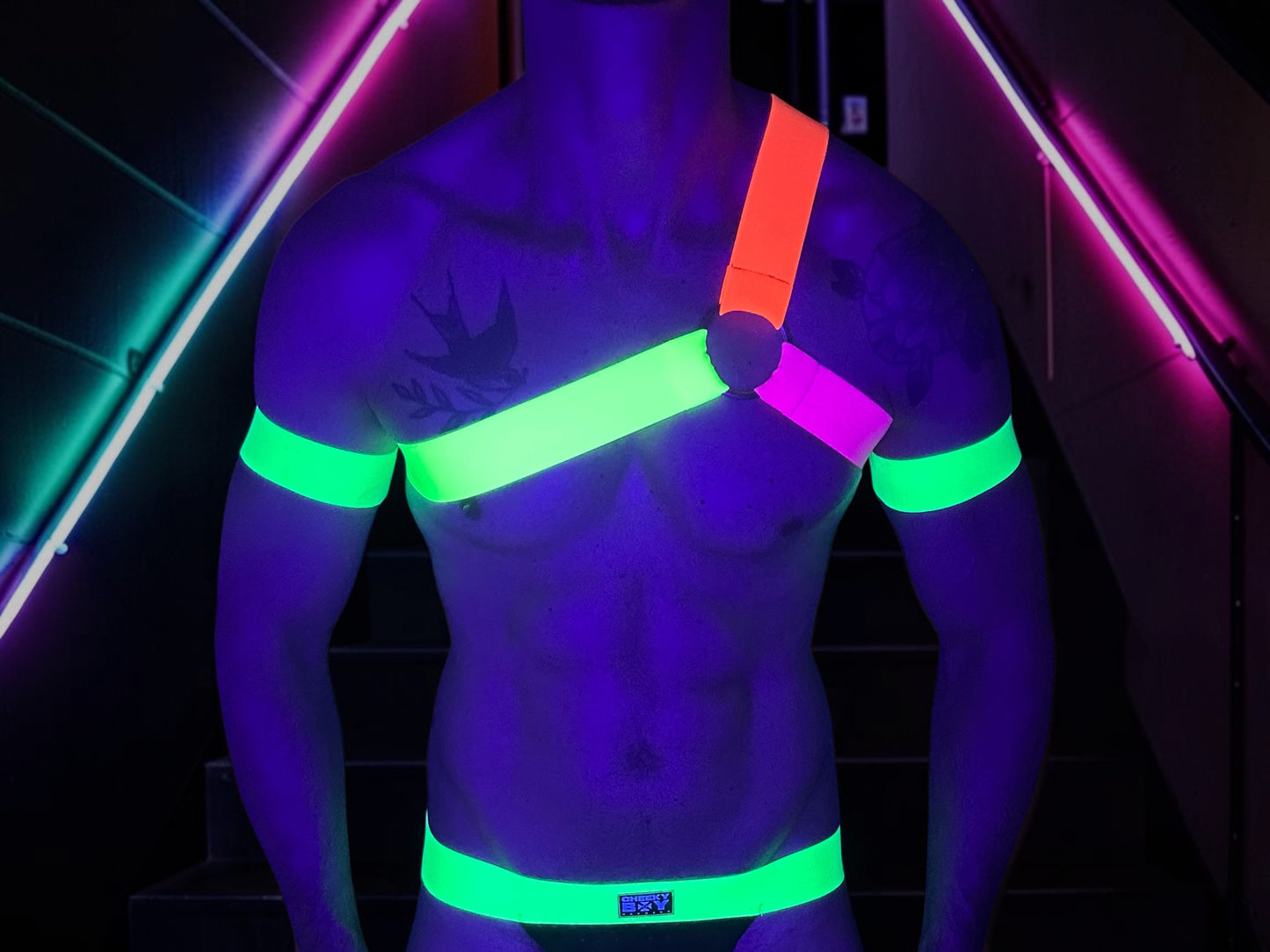 Toned male model wearing a made-to-measure Spartan-style neon chest harness with matching jockstrap, thigh bands and bicep bands in neon green, neon orange, neon pink and neon yellow elastic straps under UV lighting