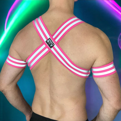 Made-to-measure men’s elastic chest harness in high-visibility neon pink with reflective detailing, glowing under blacklight. The design features double shoulder and chest straps joined by circular O-rings, with optional matching jockstrap and thigh bands—ideal for clubwear, festivals, or fetish fashion.