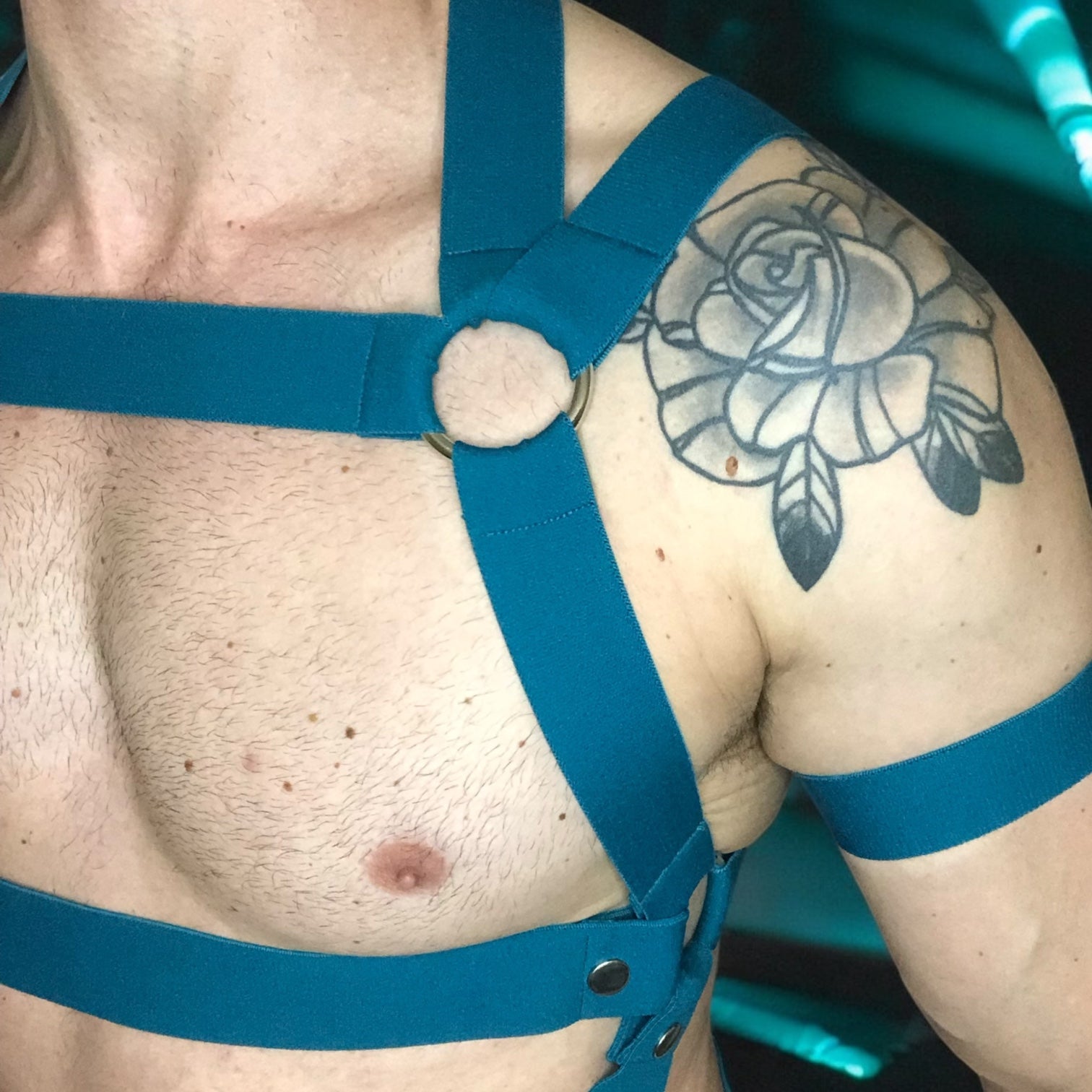 Cheeky Boy Harness, featuring geometric strap design with metal rings across the chest and torso. The outfit includes matching teal armbands and a black mesh jockstrap. Set against a futuristic, teal-lit club backdrop, perfect for festival or rave wear.