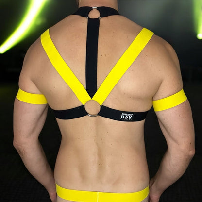 Model wearing a custom-made yellow and black elastic chest harness with metal rings and matching jockstrap by Cheeky Boy Harness. Includes red bicep bands and leg straps, designed for clubwear, fetish events, and festivals